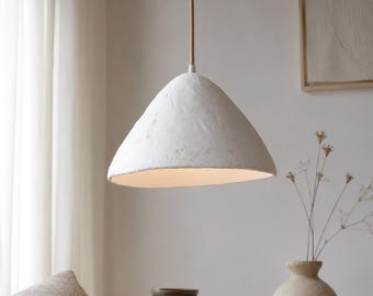 May include: A white pendant light with a textured, organic, bell-shaped shade suspended by a thin cord. The shade has a matte, uneven surface finish and a wide opening at the bottom, casting a soft light downward from the interior of the fixture.