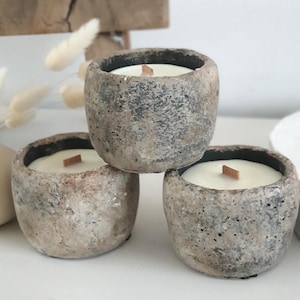 Könnte beinhalten: Drei kleine, runde Kerzen in strukturierten, grau-beigen Betonbehältern. Jede Kerze hat eine weiße Wachsfüllung und einen Holzdocht. Die Kerzen sind in einem kleinen Stapel angeordnet, wobei die oberste Kerze leicht dezentriert ist.