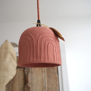 Puede incluir: Una lámpara colgante de color terracota con una pantalla en forma de campana. La pantalla presenta una serie de ranuras curvas en forma de arco. La luz cuelga de un cable marrón y tiene una pequeña etiqueta. El diseño es minimalista y moderno.