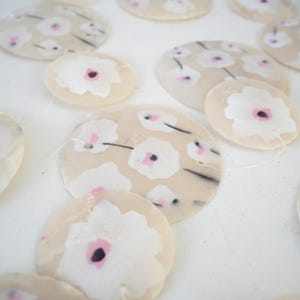 K&ouml;nnte beinhalten: Eine Ansammlung von kreisf&ouml;rmigen, durchscheinenden, cremefarbenen Scheiben mit einem sich wiederholenden Muster aus wei&szlig;en Blumen mit dunklen Mitten und rosa Akzenten. Jede Scheibe ist mit einem d&uuml;nnen, klaren Faden verbunden, was ein h&auml;ngendes Arrangement ergibt.