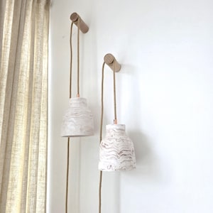 May include: Two pendant lights with a marbled white and brown pattern hang from wooden wall mounts. The lights have copper-colored accents and are suspended by brown rope cords. The background is a white wall and a beige curtain.