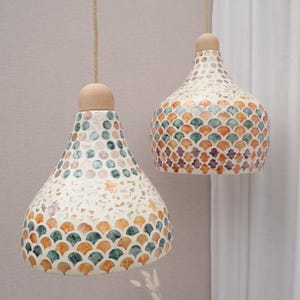 Op de afbeelding: Twee hanglampen met een mozaïekontwerp. De lampenkappen zijn wit met een patroon van oranje, blauwgroene en paarse mozaïektegels. Elke lamp heeft een houten bovenkant en is opgehangen aan een natuurlijk touw.