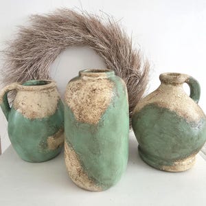 Könnte beinhalten: Drei Keramikvasen in verschiedenen Formen und Größen mit einem zweifarbigen Grün-Beige-Farbschema. Die Vasen haben ein rustikales, strukturiertes Aussehen, mit einem getrockneten Kranz im Hintergrund. Die Vasen eignen sich für die Inneneinrichtung.