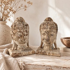 Puede incluir: Dos esculturas de cabezas de Buda talladas en madera con un acabado desgastado y envejecido en tonos beige y marrón. Cada cabeza está montada sobre una base cuadrada, mostrando rizos de cabello detallados, ojos cerrados y lóbulos alargados.