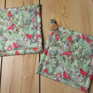 May include: Two quilted pot holders with a wintery design featuring red cardinals, holly, and snow on a green background. Each pot holder has a brown button.