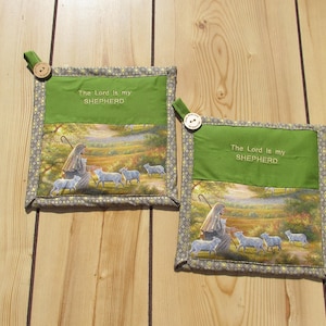 May include: Two green and gray pot holders with a floral pattern and the text "The Lord is my SHEPHERD". The pot holders feature a picture of a shepherd with sheep in a field.