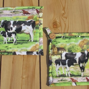 May include: Two square pot holders with a farm scene featuring cows and a calf in a green field. The pot holders have a loop with a wooden button for hanging. The edges are trimmed with a green and white patterned fabric.