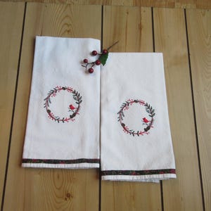 May include: Two white hand towels with a decorative embroidered wreath design featuring a red bird. The wreath is composed of green leaves, red berries, and pinecones. The towels have a patterned border.