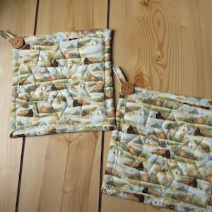 May include: Two quilted pot holders with a landscape pattern featuring barns, fields, and a blue sky. Each pot holder has a fabric loop and a wooden button for hanging. The pot holders are square and measure approximately 8 inches by 8 inches.