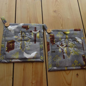May include: Two gray square pot holders with a religious theme. Each pot holder features a cross, a dove, and a book design. The pot holders have a button and loop for hanging.