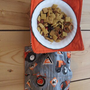 May include: A white bowl filled with a snack mix, resting on an orange quilted hot pad. Below, a gray fabric pocket features orange tractors and the "Allis-Chalmers" logo. The background is a wooden surface.