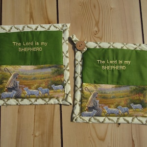 May include: Two square fabric pot holders featuring a green upper section with the embroidered text "The Lord is my SHEPHERD" and a lower section depicting a shepherd with sheep in a field. Each piece has a cream border and a wooden button.