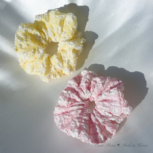 May include: Two fabric hair scrunchies, one yellow and one pink, with a ruffled texture. The scrunchies are on a white satin background.