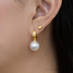 May include: A pair of gold earrings with a pearl drop and a small heart charm. The earrings are worn in a person's ear.