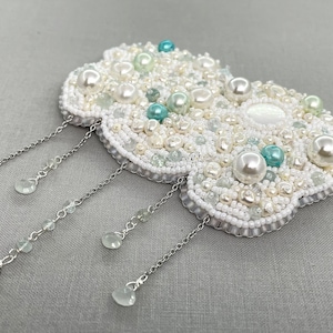 Peut inclure: Une broche en forme de nuage en perles blanches avec des perles turquoise et blanches, avec des chaînes argentées et des perles turquoise pendantes.