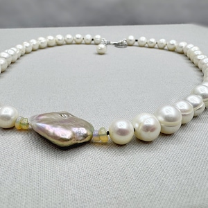 May include: A white pearl necklace with a large, irregular, iridescent pearl pendant and a smaller pearl charm. The necklace is strung on a black cord.