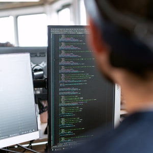 May include: A person looking at a computer screen displaying code. The code is written in a dark background with colourful syntax highlighting.