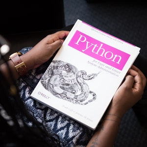 Puede incluir: Un libro blanco con un t&iacute;tulo rosa que dice "Python for Unix and Linux System Administration" de O'Reilly. El libro presenta una ilustraci&oacute;n en blanco y negro de una serpiente.