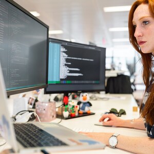 May include: A woman sits at a desk in front of a laptop and two computer monitors. The monitors display code. The woman is wearing a blue and white patterned shirt and has long red hair.