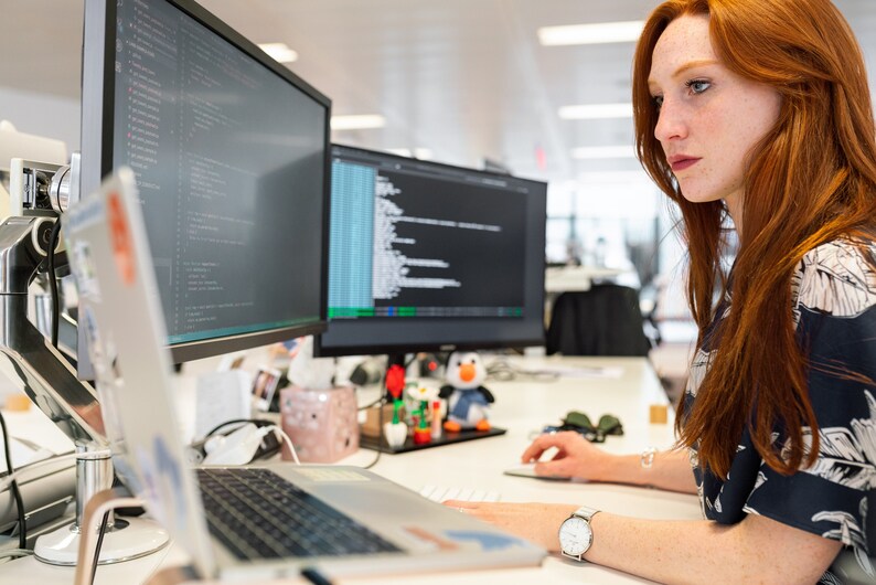May include: A woman sits at a desk in front of a laptop and two computer monitors. The monitors display code. The woman is wearing a blue and white patterned shirt and has long red hair.