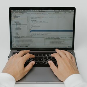 May include: A person is typing on a silver laptop computer with a black keyboard. The laptop screen displays code in a programming language.