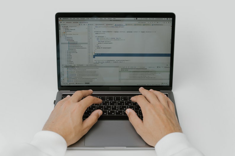 May include: A person is typing on a silver laptop computer with a black keyboard. The laptop screen displays code in a programming language.