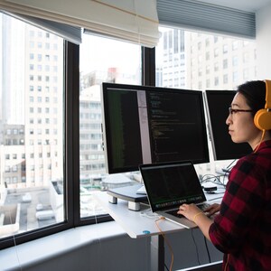 May include: A person wearing headphones sits at a desk with three computer monitors and a laptop. The monitors display code. The person is looking at the monitors and typing on the laptop.