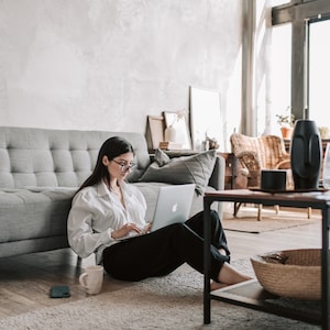 May include: A woman is sitting on the floor in front of a grey sofa, working on a laptop computer. She is wearing a white button-down shirt and black trousers. A mug and a phone are on the floor next to her.