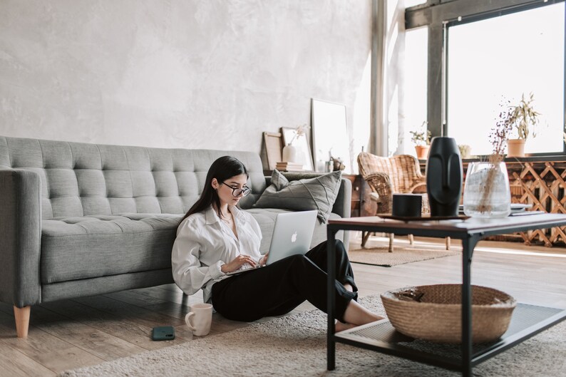 May include: A woman is sitting on the floor in front of a grey sofa, working on a laptop computer. She is wearing a white button-down shirt and black trousers. A mug and a phone are on the floor next to her.