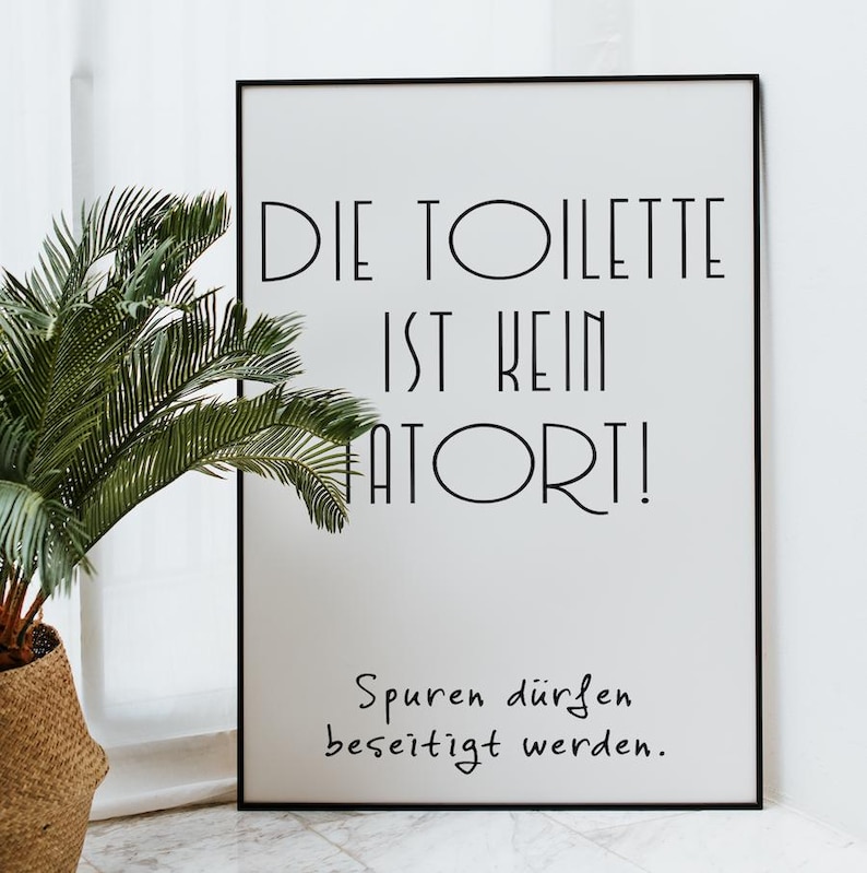 May include: A framed white print with black text that reads "DIE TOILETTE IST KEIN TATORT! Spuren d&uuml;rfen beseitigt werden." A potted plant with green fronds is in the foreground. The frame is black.