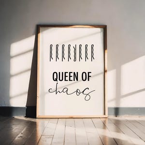 Könnte beinhalten: Ein gerahmter Druck mit dem Text "QUEEN OF chaos" in schwarzer Schrift. Über dem Text befinden sich schwarze Haarnadeln. Der Rahmen ist aus Holz. Der Druck lehnt an einer weißen Wand auf einem Holzboden.