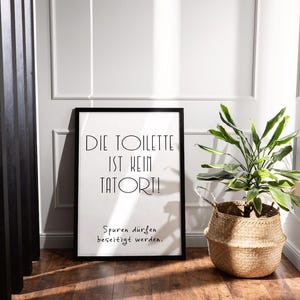 May include: A framed print with the text "DIE TOILETTE IST KEIN TATORT!" in black, with the phrase "Spuren d&uuml;rfen beseitigt werden." below it. A woven basket with a green plant sits to the right. The room has wood flooring and white walls.