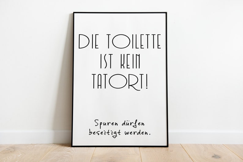 May include: A framed poster with black text on a white background. The text reads "DIE TOILETTE IST KEIN TATORT!" and "Spuren d&uuml;rfen beseitigt werden." The poster is in a black frame and leans against a white wall.