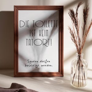 May include: A framed print with the text "DIE TOILETTE IST KEIN TATORT!" in black, with the phrase "Spuren d&uuml;rfen beseitigt werden." below. The frame is brown, and a clear glass vase with dried plants sits to the right.
