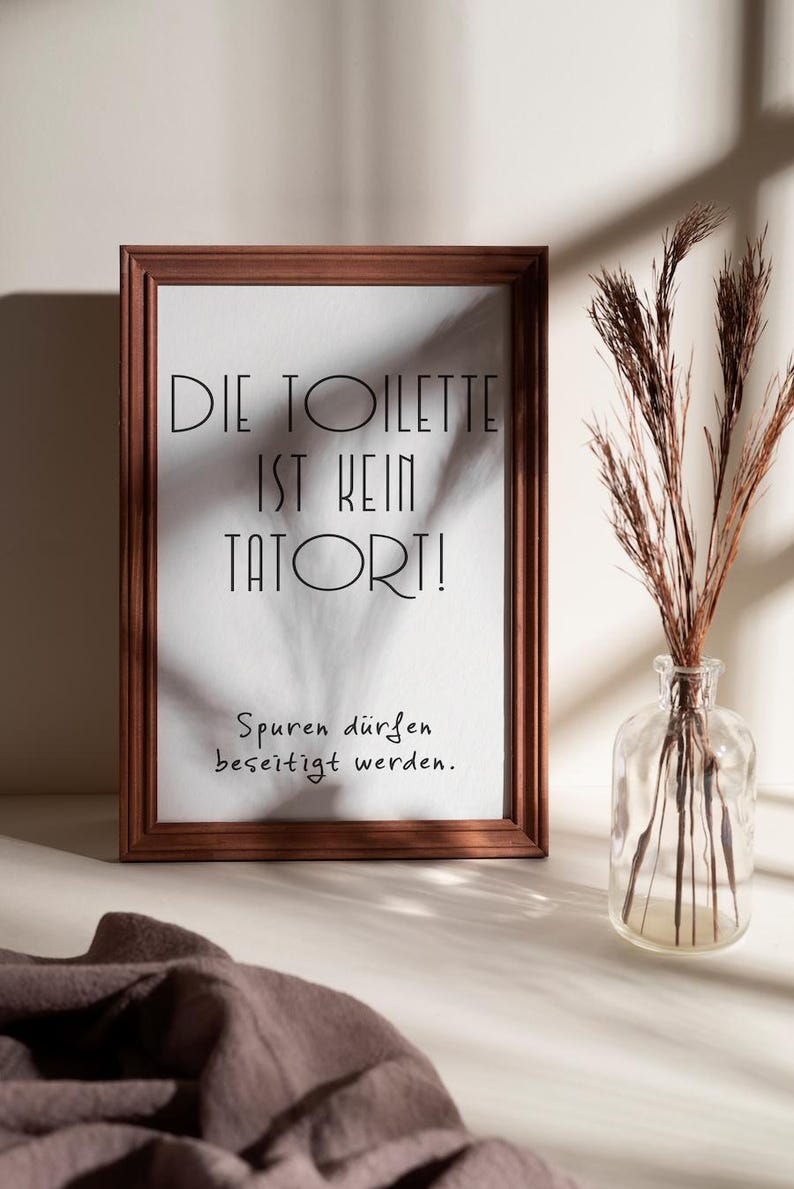 May include: A framed print with the text "DIE TOILETTE IST KEIN TATORT!" in black, with the phrase "Spuren d&uuml;rfen beseitigt werden." below. The frame is brown, and a clear glass vase with dried plants sits to the right.