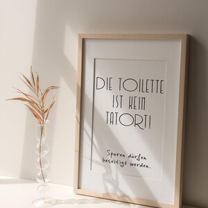 May include: A framed print with the text "DIE TOILETTE IST KEIN TATORT!" in black, with the phrase "Spuren d&uuml;rfen beseitigt werden." below. A clear glass vase with a dried plant is to the left.