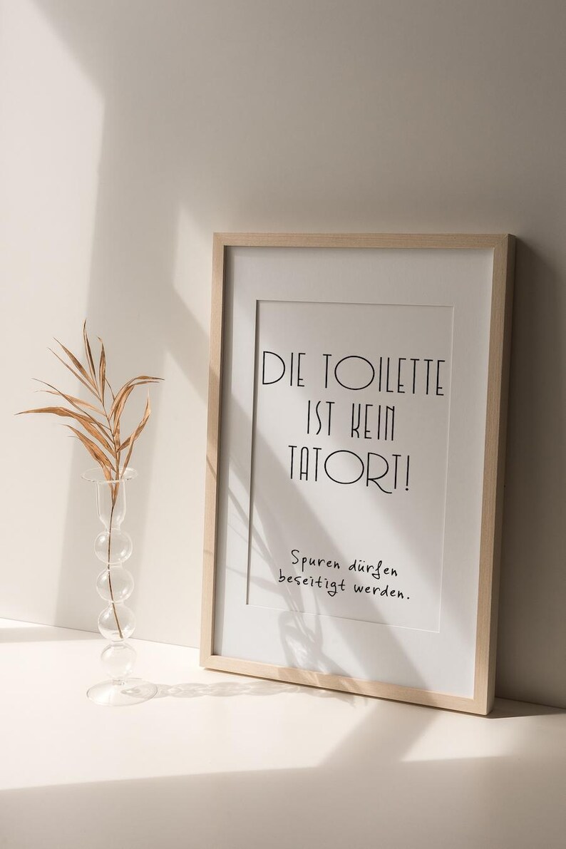 May include: A framed print with the text "DIE TOILETTE IST KEIN TATORT!" in black, with the phrase "Spuren d&uuml;rfen beseitigt werden." below. A clear glass vase with a dried plant is to the left.