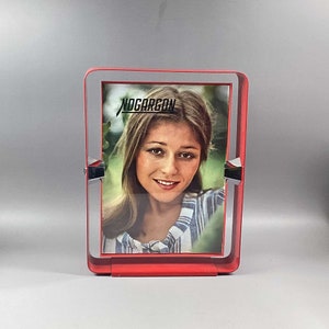 Puede incluir: Un marco de fotos de metal rojo con un borde plateado y una foto de una mujer en el interior. El marco tiene la palabra "Nogarcon" impresa en la parte superior.