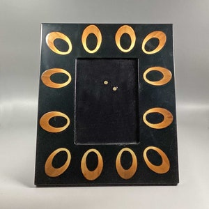 Puede incluir: Un marco de fotos negro con un diseño ovalado dorado alrededor del borde. El marco tiene un paspartú negro y un cristal transparente en la parte delantera.