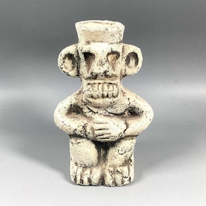 May include: White ceramic figurine of a seated figure with a wide, toothy grin and large ears. The figure has a rounded, almost bulbous head and a simple, stylized body.