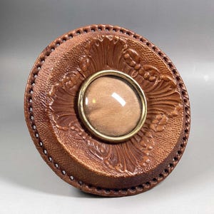 May include: A round, brown leather box with a decorative floral pattern and a gold-colored metal frame. The box has a clear, circular glass insert in the center. The edges of the box are stitched.