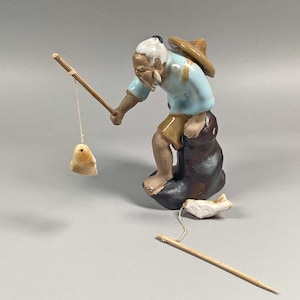 May include: A ceramic figurine of a fisherman with a blue shirt, brown pants, and a straw hat. He is holding a fishing rod and a small fish is attached to the line. The figurine is sitting on a brown rock.