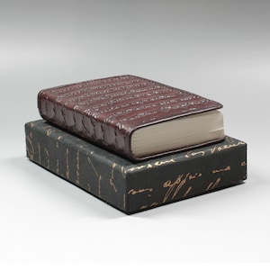 Könnte beinhalten: Ein braunes, ledergebundenes Tagebuch mit einer strukturierten Oberfläche liegt auf einer Schachtel mit goldener Schrift.