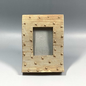 Puede incluir: Un marco de fotos dorado con una superficie texturizada y un patrón de pequeños puntos dorados. El marco tiene una abertura rectangular para una foto.