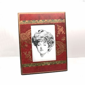 Puede incluir: Un marco de fotos rojo con un diseño de cachemira dorado y verde. El marco tiene una imagen en blanco y negro de una mujer con el pelo rizado.
