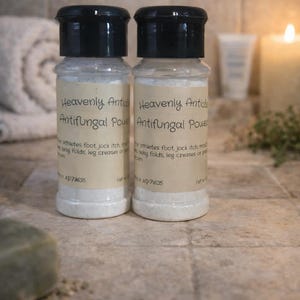 May include: Two clear plastic bottles with black screw-top lids containing white powder. Each bottle features a beige label with the text Heavenly Antifungal Powder, listing uses for athlete's foot, jock itch, and skin folds, with code AP71635.