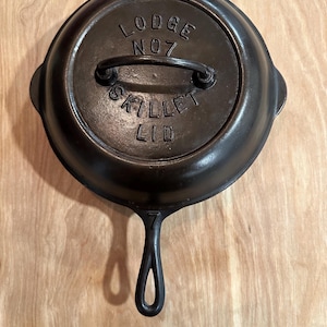 May include: A black cast iron skillet with a matching lid. The lid is embossed with the text "LODGE NO 7 SKILLET LID". The skillet has a handle and two small side handles. It is displayed on a light brown wooden surface.