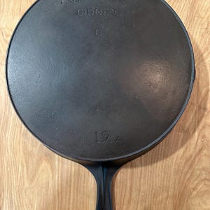 May include: A black cast iron skillet with a long handle. The skillet has the text "WAGNER SIDNEY" and "12A" embossed on the bottom. The pan is round and appears to be well-seasoned.