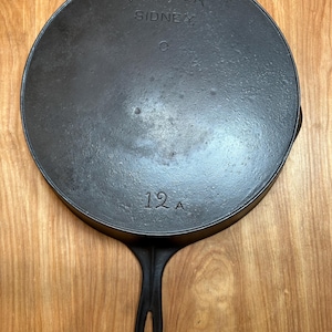May include: A black cast iron skillet with a long handle. The skillet has the text "WAGNER SIDNEY" and "12 A" embossed on the bottom. The skillet is round and appears well-seasoned.