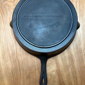 May include: A black cast iron skillet with a long handle and a circular cooking surface. The skillet is shown from above, resting on a wooden surface. The handle has a hole for hanging. The skillet is approximately 10 inches in diameter.
